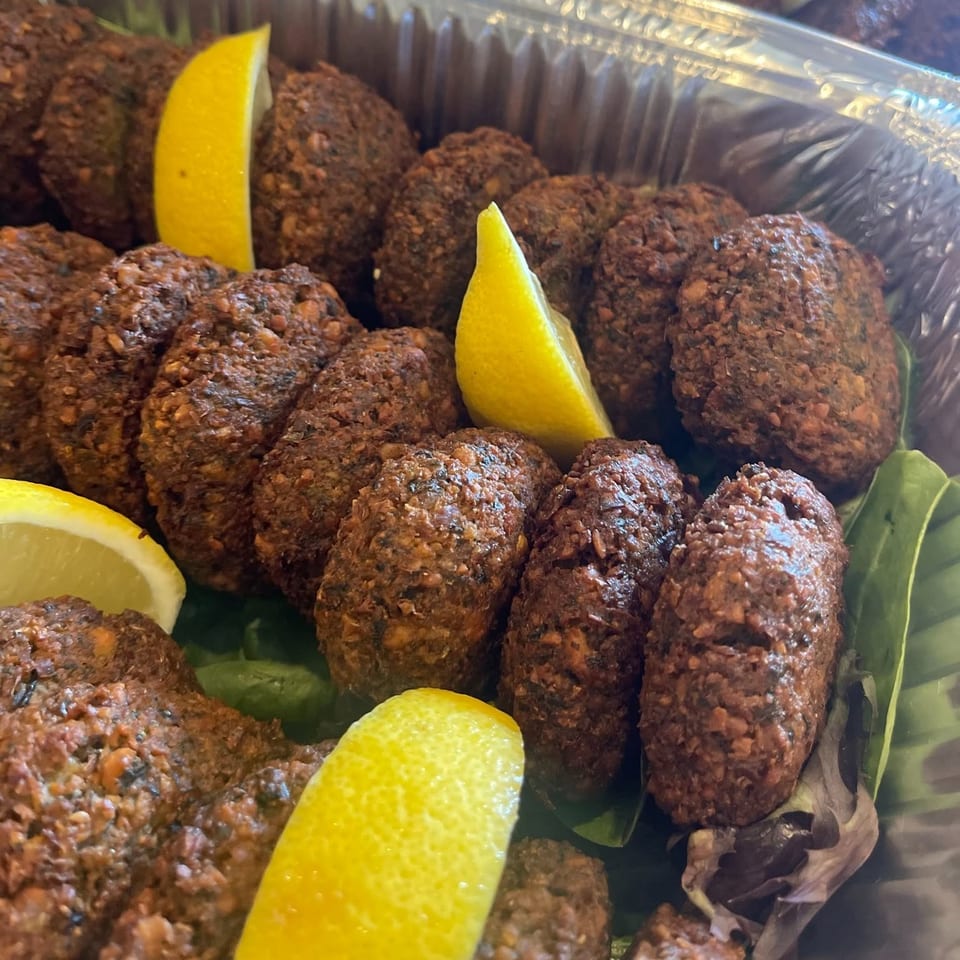 Numerous fried falafel patties with lemon wedges in a foil container on green leaves.