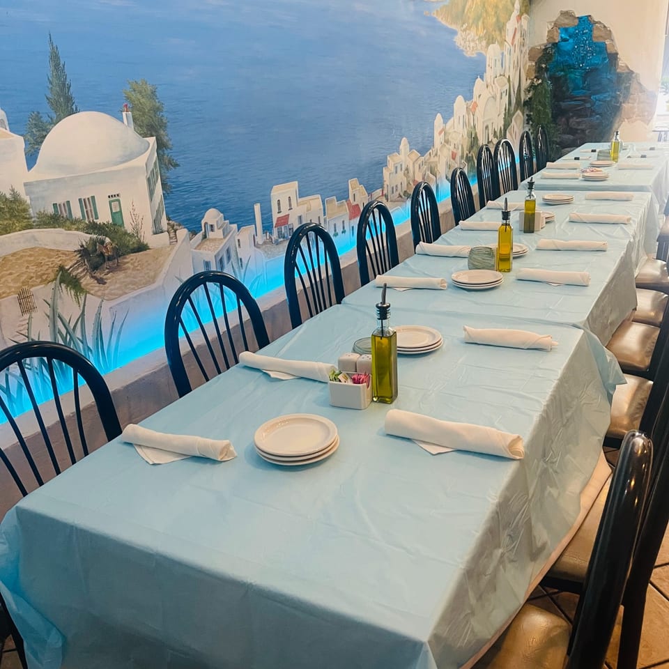 The Santorini coastline mural behind a long table set for guests at Anna's Greek Cuisine