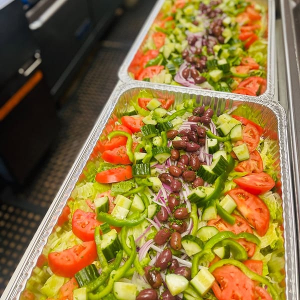 Two large aluminum trays filled with fresh salads, featuring lettuce, tomatoes, cucumbers, green peppers, red onions, and olives.