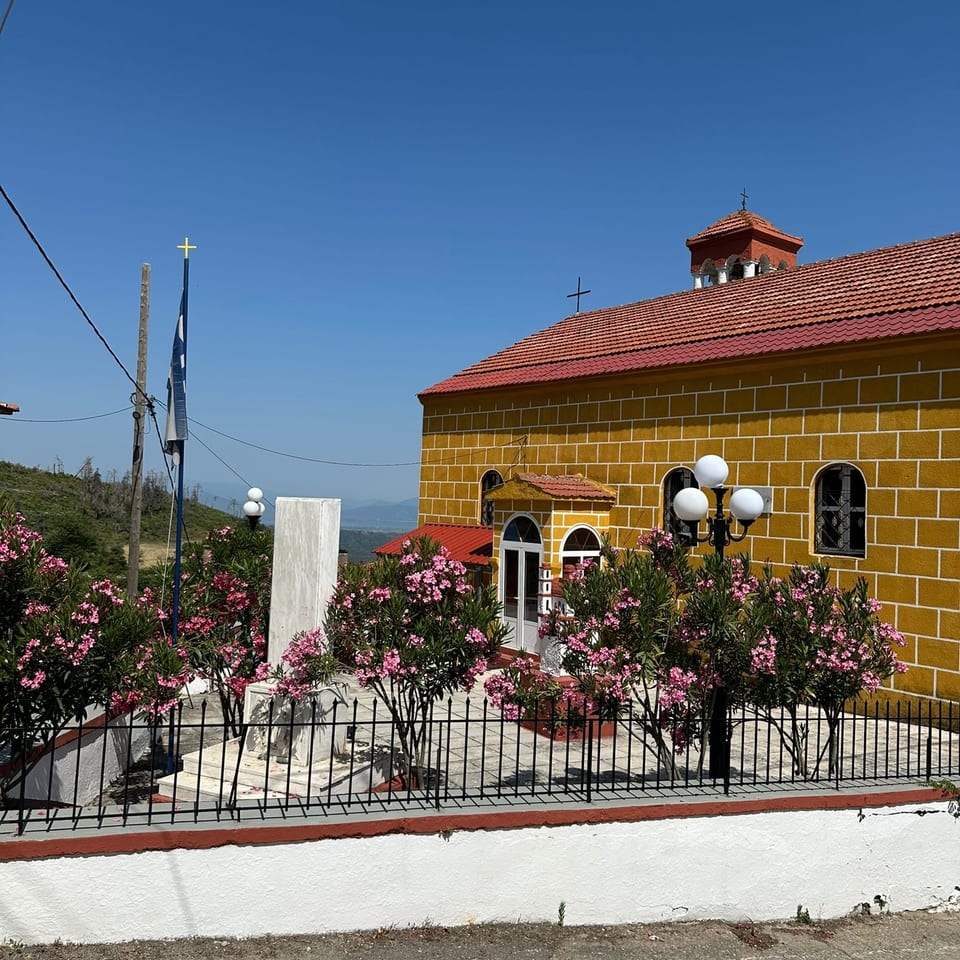 Yellow stone church with red tile roof, crosses, flowering shrubs, and mountain landscape under a clear blue sky.